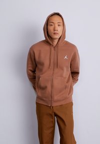 Sudadera con cremallera marrón, con un bolsillo canguro frontal y el logo de Jordan en blanco. Presenta una capucha con cordones, puños acanalados y una textura suave.