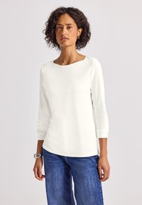 Haut blanc à manches longues avec encolure bateau et coupe slim, associé à un jean bleu à jambes larges. Texture lisse, design minimal, style décontracté.