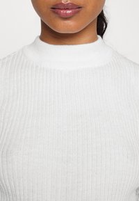 Pull à col roulé blanc en maille avec une texture côtelée verticale, doté d'un col montant. Matière douce avec une silhouette ajustée.