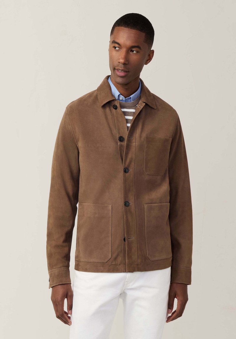 Giacca in suede marrone con quattro tasche, chiusura a bottone e colletto. Indossata sopra un maglione a righe e pantaloni bianchi. Tessuto liscio.