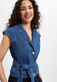 Ärmlös blå denimvest med krage, två knappar och bälte i midjan. Har synlig sömnad och en strukturerad passform.