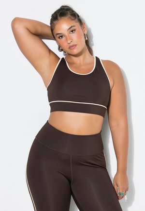 Vrouw die een donkerbruine sportbeha en een legging met lichte bies draagt, poseert met één arm omhoog tegen een witte achtergrond.