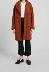 Orange imitation pälsjacka med bred krage, knäppning framtill och avslappnad passform, i kombination med svarta rutig kortbyxor och svarta loafers.