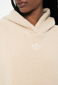Krémová mikina s kapucňou z fleece s textúrovaným povrchom, prednou vreckom a prišitým logom blízko výstrihu.