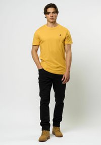 Tricou din bumbac galben cu guler rotund și un mic logo, asortat cu blugi negri și bocanci maro cu șireturi. Design simplu, ajustat.