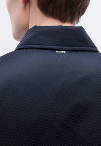 Μπλε ναυτικό υφασμάτινο σακάκι με λεπτό ρόμβο σχέδιο και μυτερό γιακά. Διαθέτει ασημένια ετικέτα με λογότυπο στο πίσω μέρος του λαιμού.