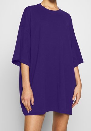 Femme portant une robe violette à manches courtes surdimensionnée avec un col rond, se tenant devant un fond blanc uni.