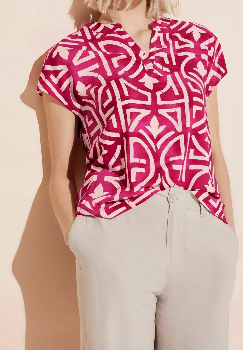 Blouse à manches courtes avec col en V, de couleur rose vif avec des motifs géométriques blancs. Fabriquée en tissu léger, elle présente une coupe décontractée.