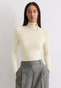 Vrouw met lang donker haar, gekleed in een crèmekleurige coltrui en hooggesneden grijze broek, met handen in de zakken tegen een effen achtergrond.