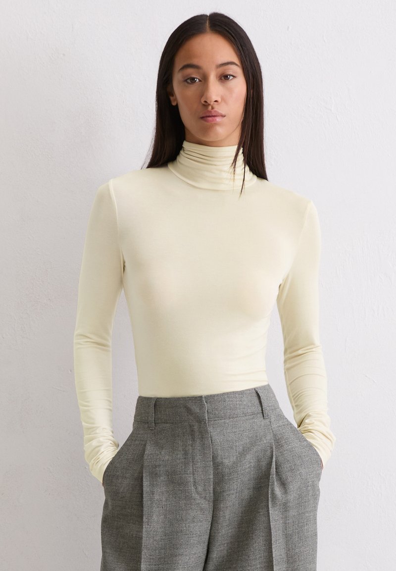 Vrouw met lang donker haar, gekleed in een crèmekleurige coltrui en hooggesneden grijze broek, met handen in de zakken tegen een effen achtergrond.