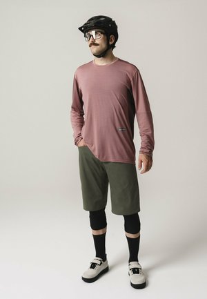 Mann trägt einen schwarzen Fahrradhelm, eine Brille, ein mauvefarbenes Langarmshirt, olivgrüne Shorts, schwarze Knieschoner und weiße Turnschuhe und steht.