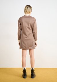 Braunes Baumwollkleid mit langen Ärmeln und cremefarbenen Bündchen, mit gerader Schnittführung und minimalem Design. Schwarze Knöchelschuhe mit dicker Sohle.