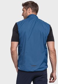 Blauwest mit leichtem Gewicht, glatter, matter Textur, hohem Kragen und akzentuierender reflektierender Partie am unteren Rücken. Getragen über einem schwarzen Shirt.
