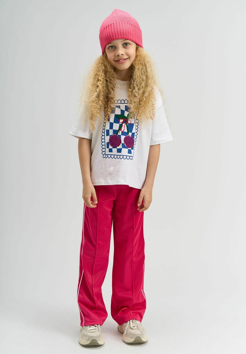 Enfant portant un t-shirt blanc avec un motif à carreaux coloré et un graphisme de cerises, associé à un pantalon de survêtement rose vif et des baskets.
