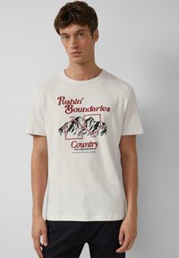 Młody mężczyzna ubrany w białą koszulkę z czerwoną i czarną grafiką gór oraz napisem "Pushin' Boundaries Country Pacemakers" na jednolitym tle.
