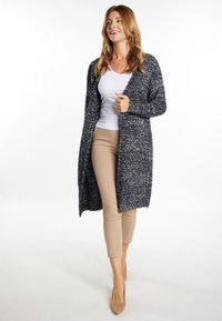 Langer, offenfrontiger Cardigan aus einem schwarz-weißen Text Strick. Kombiniert mit einem weißen Oberteil und beigen, engen Hosen. Neutrale hohe Absätze runden das Outfit ab.