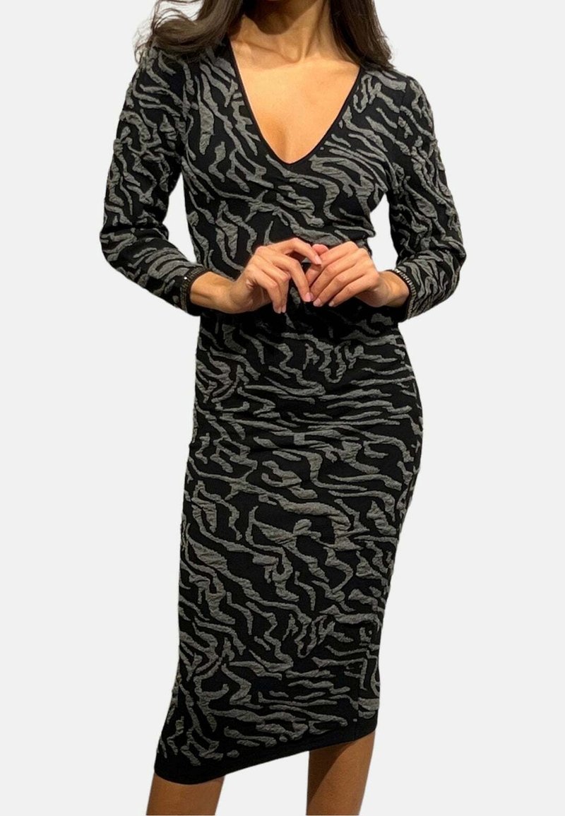 Femme portant une robe midi noire à manches longues avec des rayures zébrées abstraites grises et un col en V, debout sur un fond blanc.