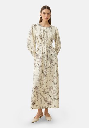 Kvinde iført en lang cremefarvet kjole med sorte blomsterprint, lange ærmer, et bælte i taljen og beige lavhælede sko, stående på en hvid baggrund.