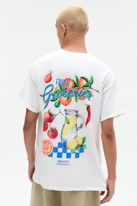 White T-shirt featuring colorful fruit and beverage graphics. Text says "FRESH Groceries." Design includes tomatoes, citrus, and a pitcher.