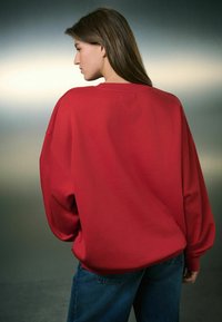 Hanorac roșu supradimensionat cu umeri lăsați, manșete ribbate și o textură netedă, combinat cu blugi din denim albastru. Vedere din spate.