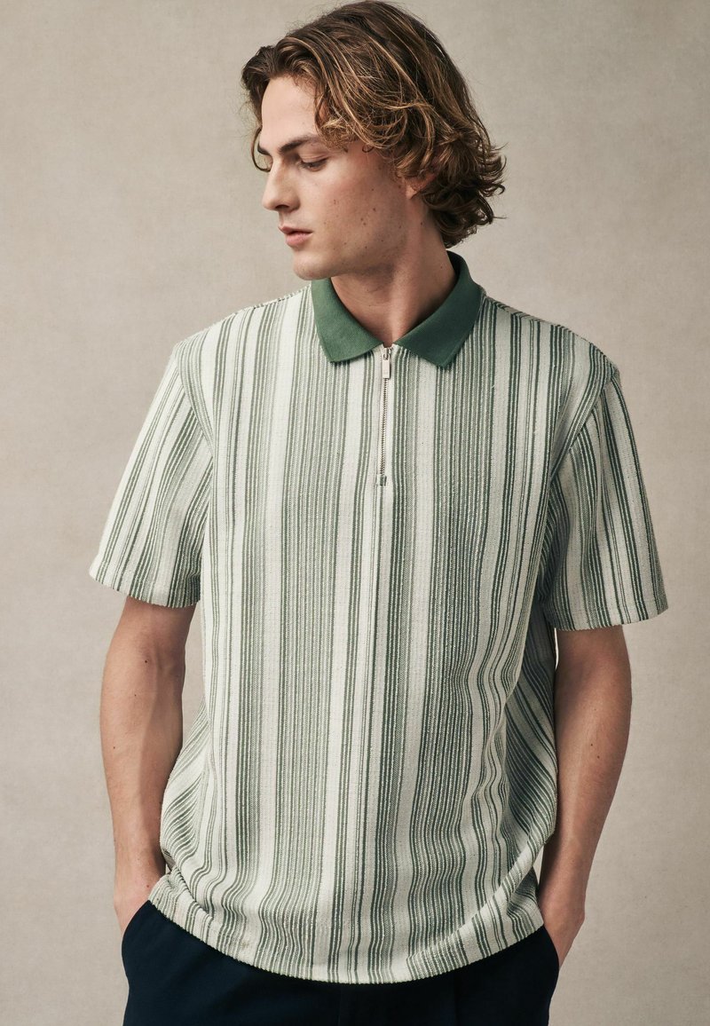 Stribet polo shirt i grønne og hvide farver, kortærmet, med lynlås og en grøn krave. Blød tekstur, afslappet pasform.