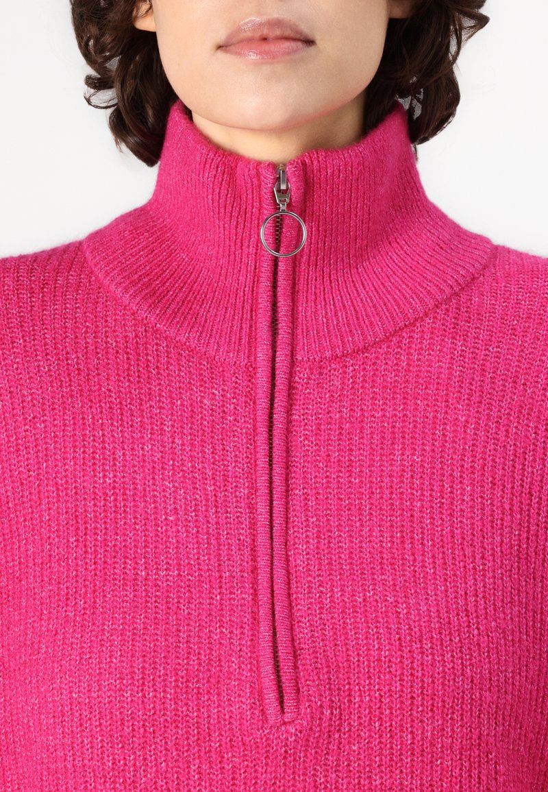 Fuksjowy sweter z dzianiny z wysokim kołnierzem, z zamkiem błyskawicznym z przodu kończącym się okrągłym uchwytem. Tekstura ma żebrowany wzór, co nadaje mu przytulny wygląd.