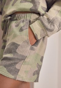 Szorty z nadrukiem kamuflażowym w stonowanych odcieniach zieleni i szarości. Posiadają elastyczny pasek w talii oraz boczną kieszeń. Wykonane z miękkiego, strukturalnego materiału.