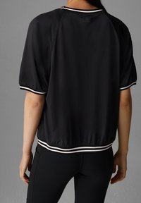 Černý krátký top s texturovaným povrchem, zdobený bílými pruhovanými akcenty na rukávech a lemu. Volný střih, lehký materiál.