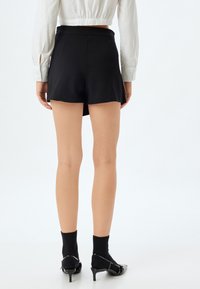 Schwarze Shorts mit einer taillierten Passform, ausgestattet mit einer glatten Textur und einem ausgestellten Saum. Getragen mit schwarzen Socken und Sandalen mit Absätzen.