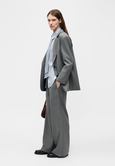 Femme vue de profil portant un costume gris surdimensionné, une chemise à rayures bleues, des chaussures noires, et tenant un sac à main marron sur un fond uni.