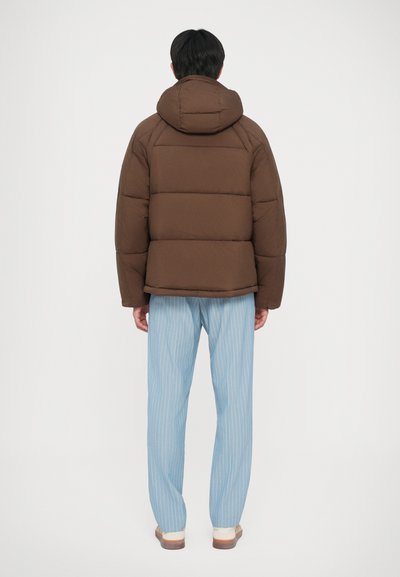 Giacca a vento marrone con cappuccio, caratterizzata da un design trapuntato. Abbinata a pantaloni a strisce blu chiaro e scarpe beige.