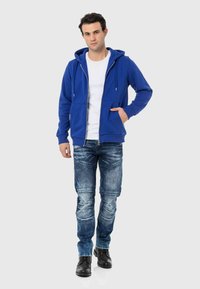 Blauwe hoodie met rits en trekkoorden, voorzakken, gedragen over een witte T-shirt; gecombineerd met versleten blauwe jeans en zwarte schoenen.