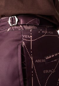 Pantalones de tela morada con un diseño de mapa en rosa claro. Detallados con una hebilla metálica y un bolsillo lateral. Textura suave.