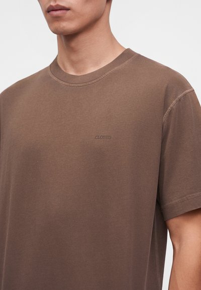 Κοντινό πλάνο ενός ατόμου που φοράει καφέ t-shirt με στρογγυλή λαιμόκοψη και τη λέξη "CLOSED" κεντημένη με μικρά γράμματα στο αριστερό στήθος.