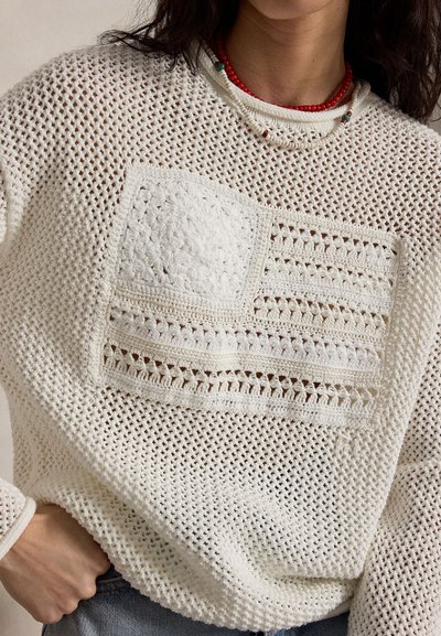 Γυναίκα που φοράει ένα λευκό πλεκτό πουλόβερ με ανάγλυφο σχέδιο αμερικανικής σημαίας στην μπροστινή πλευρά, συνδυασμένο με στρωμένα κολιέ από χάντρες.