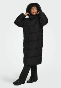 Melnā pūdera jaka ar garu dizainu, augstu apkakli, rāvējslēdzēja un pogu aizdari, kā arī šūtu tekstūru. Valgota kopā ar melnām biksēm un apaviem.