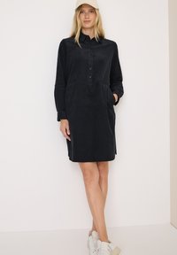 Schwarzes Cordhemdkleid mit durchgehender Knopfleiste und seitlichen Taschen. Lange Ärmel bis zu den Ellenbogen hochgerollt. Getragen mit weißen Turnschuhen und einer beigen Mütze.