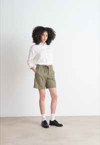 Camisa blanca de botones, pantalones cortos verde oliva con cordón, calcetines blancos hasta la rodilla y zapatos negros, de pie frente a un fondo liso.