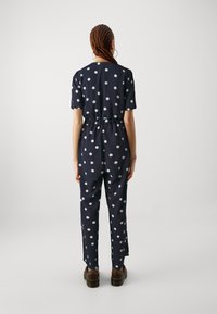 Navy jumpsuit with short sleeves, elastic waist, large white polka dots, and side slits, paired with chunky brown shoes.