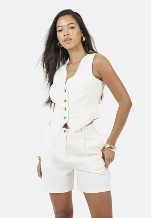 Kobieta o długich ciemnych włosach, ubrana w białą kamizelkę bez rękawów zapinaną na guziki oraz białe szorty z wysokim stanem, uzupełniona złotymi kolczykami kołami i bransoletką.