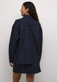 Temna denim jakna s srebrnimi bleščicami vzdolž ramen in zadnjega šiva, dolge rokave in sproščen kroj.