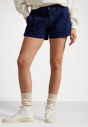 Vrouw draagt marineblauwe short, crèmekleurige trui, crèmekleurige opgekrulde sokken en witte hoge canvas sneakers, staat met handen in zakken.
