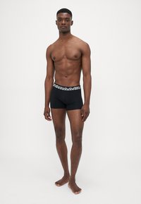 Schwarze Boxershorts mit einem weißen gemusterten elastischen Bund, aus glattem Stoff, eng geschnitten, getragen von einem oberkörperfreien männlichen Model.