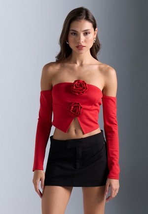 Haut court rouge à épaules dénudées avec des manches ajustées et deux embellissements en rose tridimensionnel au centre. Associé à une jupe noire.
