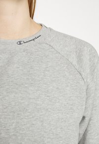 Felpa grigia chiaro con collo a giro, maniche raglan e il logo "Champion" ricamato in blu scuro sul colletto. Texture morbida.