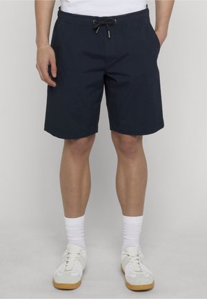 Mann trägt marineblaue Shorts mit Kordelzug, weiße Socken und weiße Turnschuhe, steht mit den Händen in den Taschen vor einem schlichten Hintergrund.