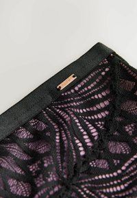 Tessuto in pizzo nero con accenti viola, caratterizzato da un motivo ondulato e bordi scallopati. Completato con un piccolo logo metallico.