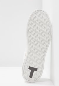 Semelle blanche de basket avec un motif géométrique texturé et un grand logo sombre en forme de T près du talon, présenté sur un fond uni.