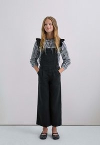 Salopette nere con gambe larghe, indossata sopra una blusa a scacchi bianca e nera con maniche a volant. Flats neri con decorazioni.