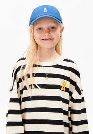 Cappello blu con logo bianco, maglione a righe bianche e nere con un dettaglio ricamato giallo. Collana con un piccolo ciondolo a forma di cuore.